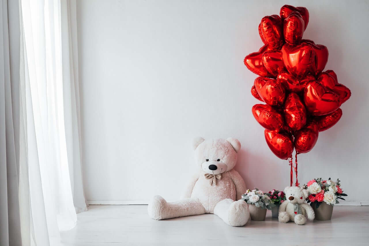 A cozy setup featuring a large white teddy bear, a bunch of shiny red heart-shaped balloons, and flower arrangements in neutral-toned pots, placed against a minimalist white background with sheer curtains.