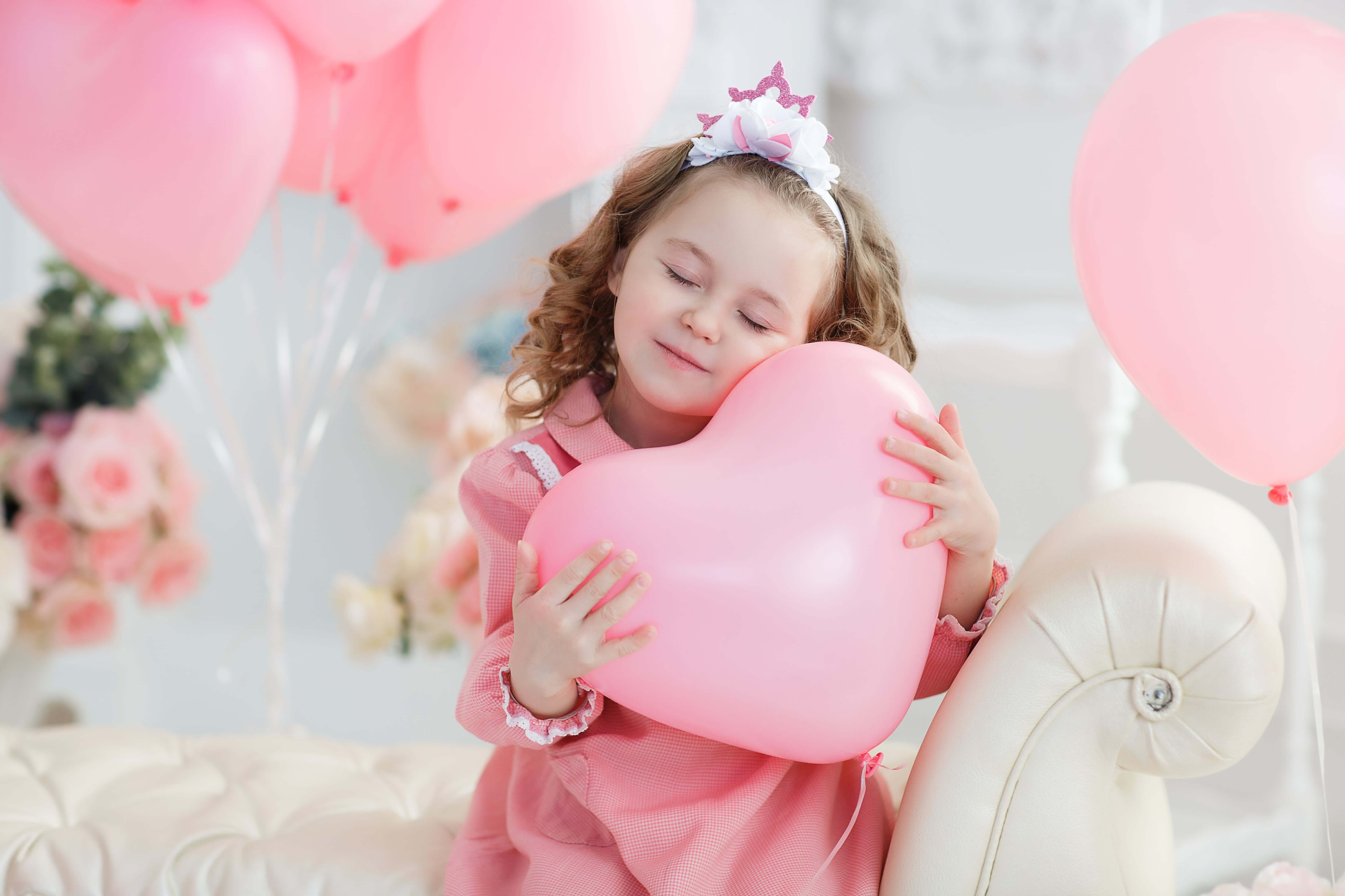 Little girl hugging a heart-shaped pink balloon with pink helium balloons in the background, perfect for kids' birthday celebrations.