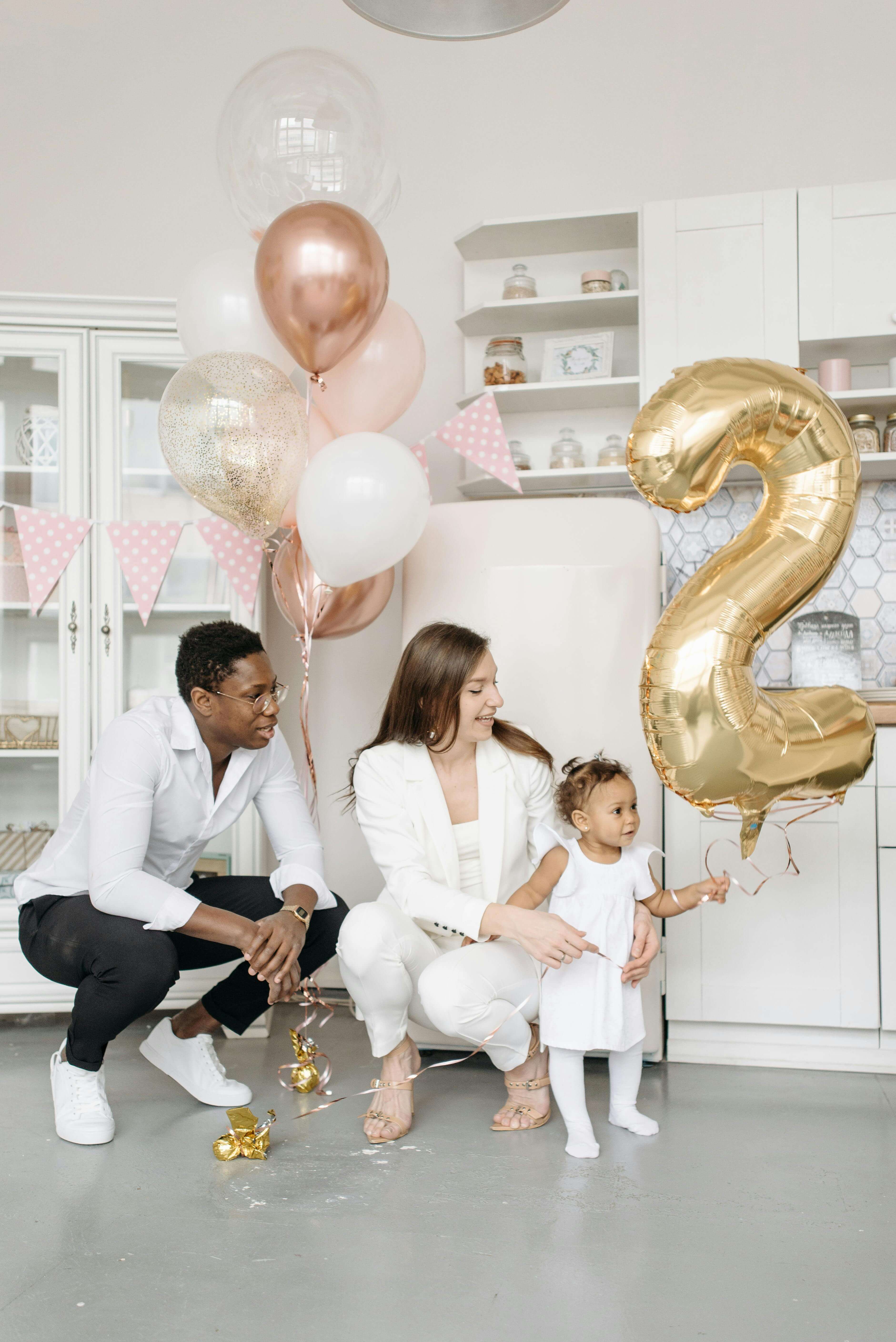 Family celebrating a child's second birthday with gold number 2 balloon, pink and white balloons, and festive decorations in a modern kitchen.