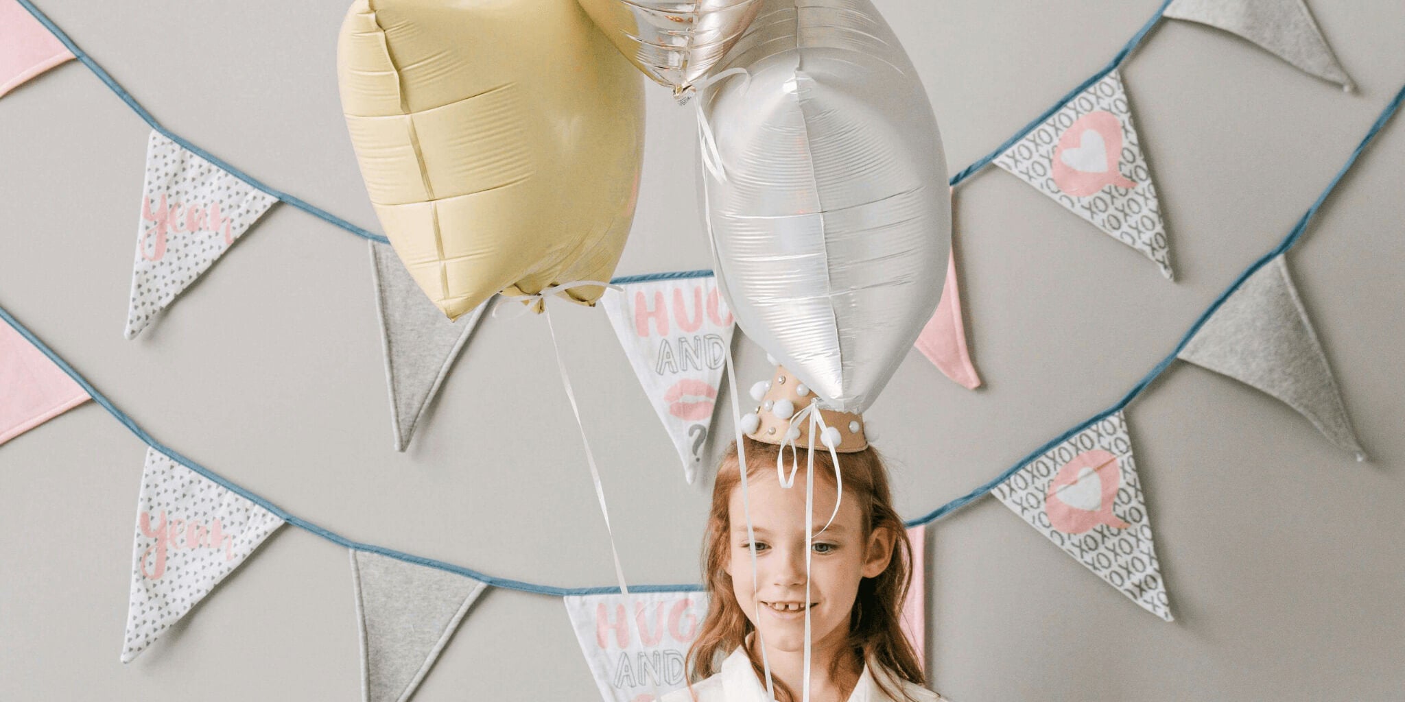 Smiling young girl wearing a party hat, holding gold and silver balloons in front of festive bunting decorations.