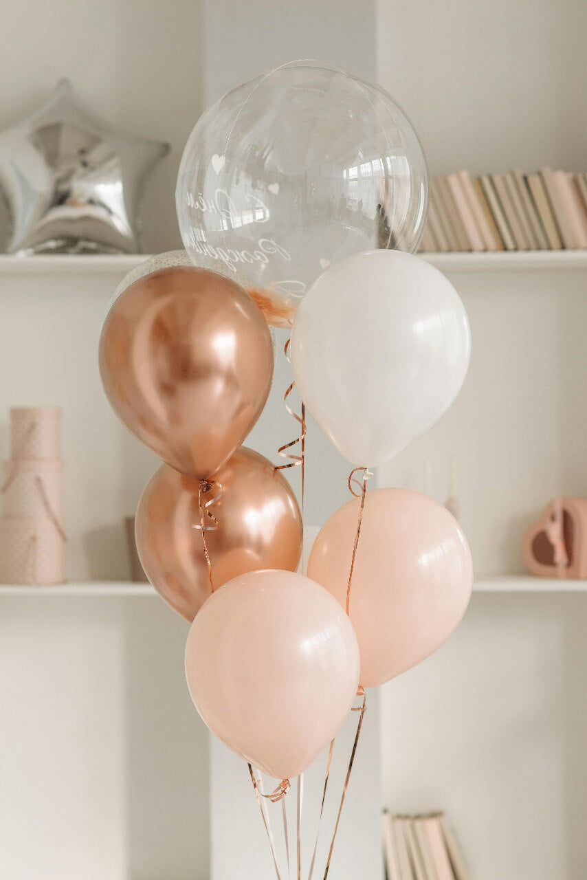 Elegant arrangement of rose gold, white, and clear balloons in front of a modern shelf with decorative items and books.