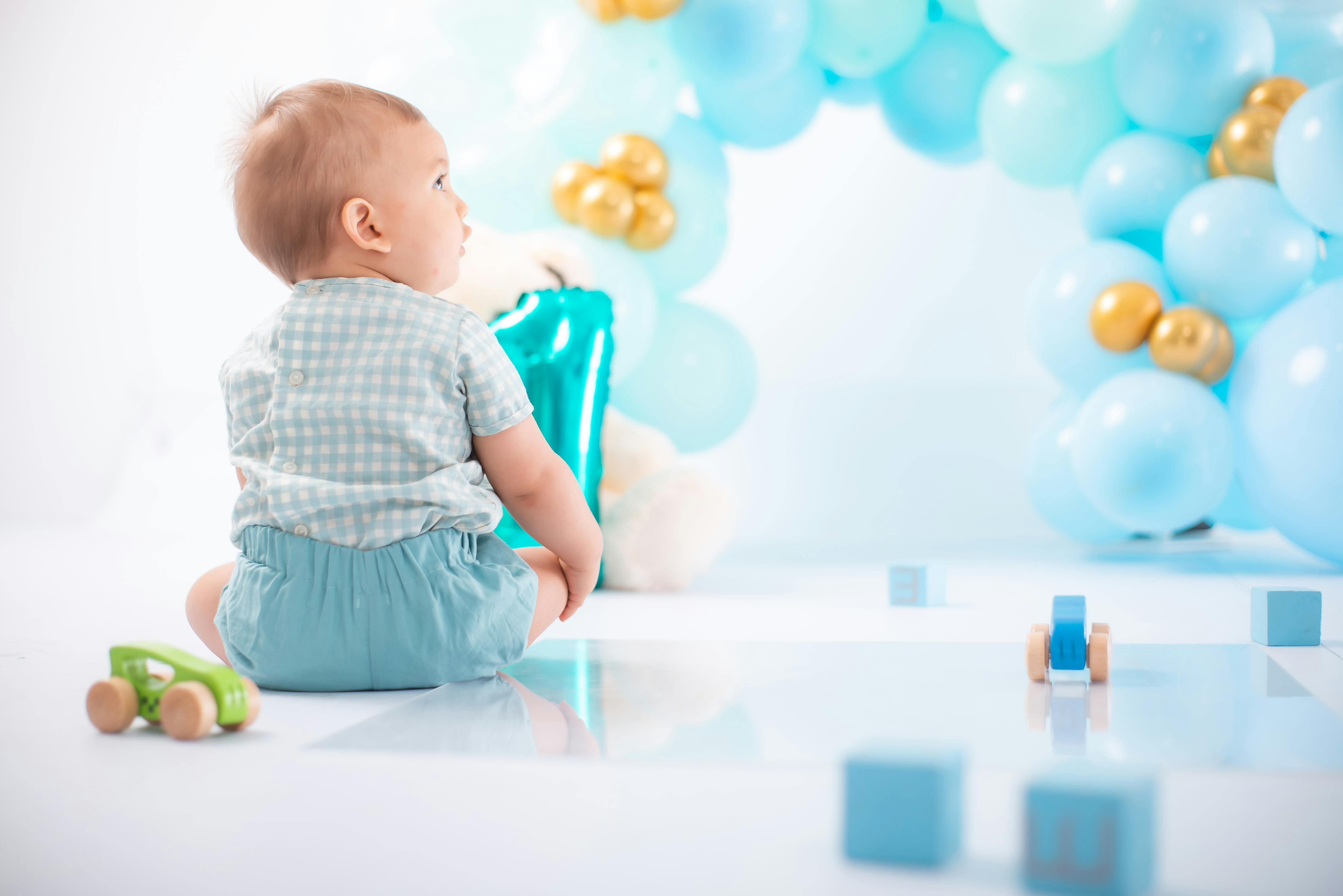 Baby in a blue outfit sits near pastel blue and gold balloons with wooden toys scattered around, suggesting a first birthday celebration.