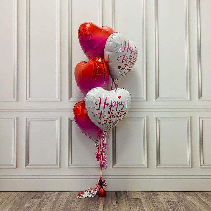 Valentine's Day balloon bouquet with red and pink ombre heart-shaped foil balloons and white heart-shaped foil balloons reading "Happy Valentine's Day," tied with pink curling ribbons and anchored with a red balloon weight.