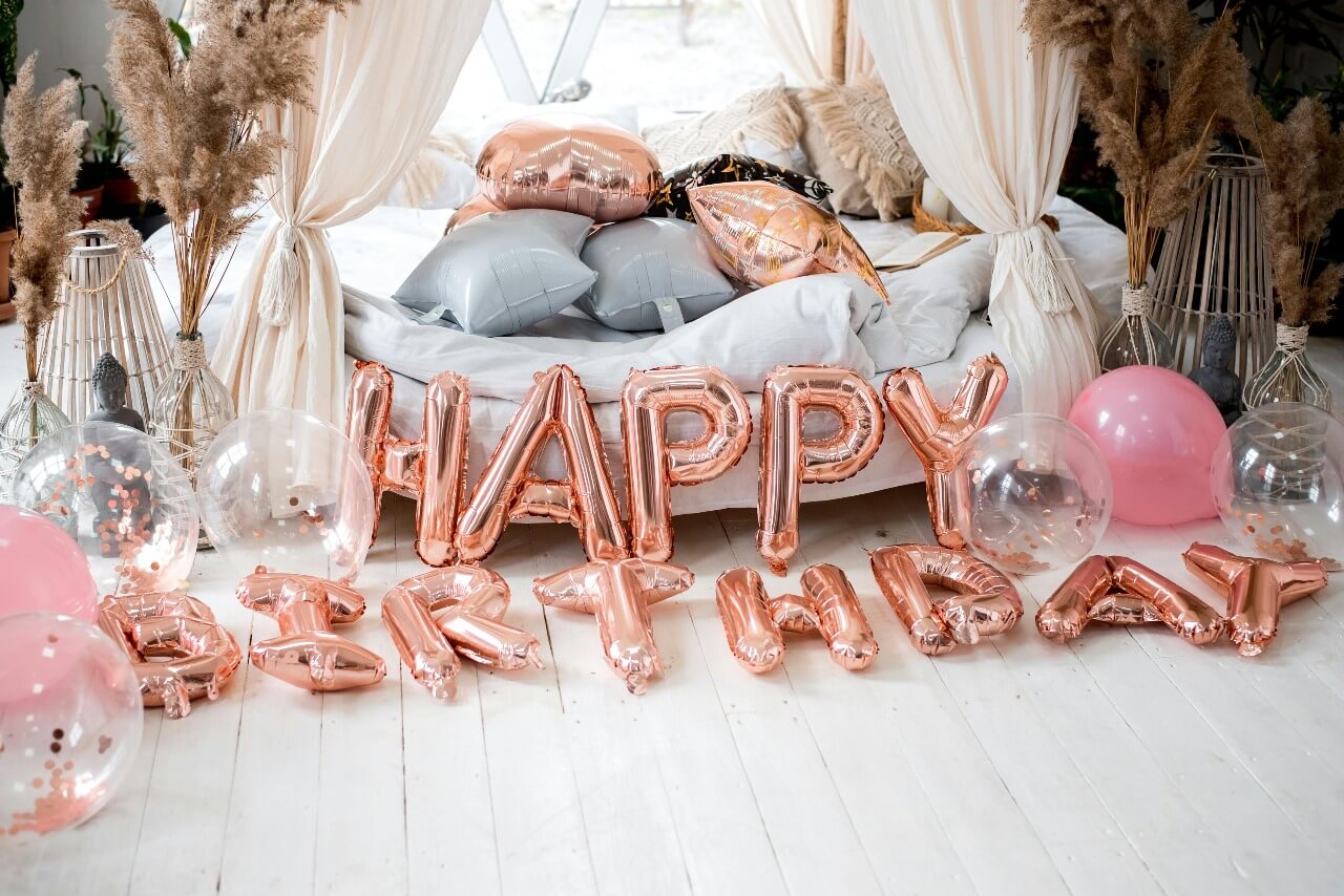 A cozy birthday setup featuring rose gold "HAPPY BIRTHDAY" foil balloons, clear and pink balloons with confetti, a white bed with decorative pillows, and pampas grass arrangements in a boho-style room.