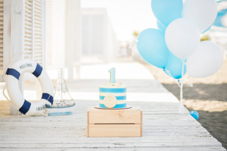 A nautical-themed first birthday setup featuring a blue and white striped cake with a number "1" topper, placed on a wooden crate. In the background, there are white and blue balloons, a lifebuoy, and a decorative sailboat, set on a wooden boardwalk by the beach.